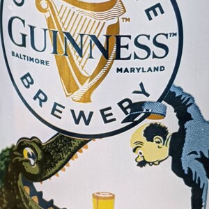 May include: A Guinness Brewery advertisement featuring the Guinness logo with a harp and the text "Baltimore Maryland". The image includes a cartoon of a man and an alligator with a glass of beer.