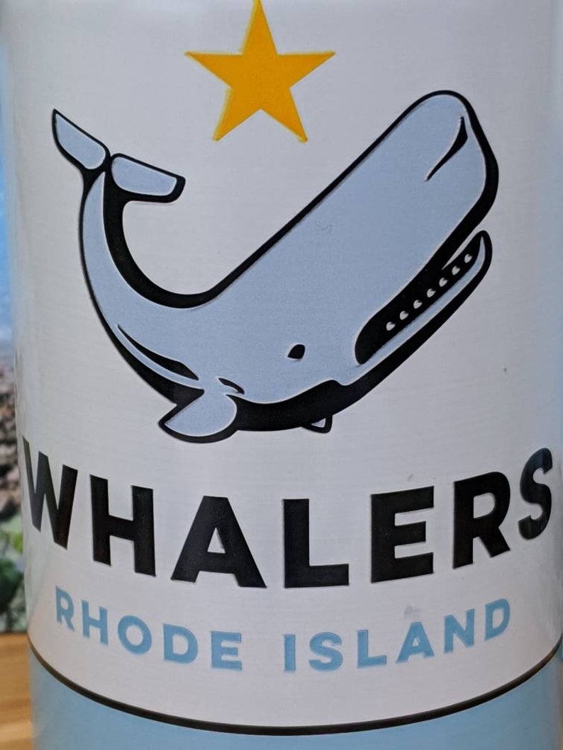 May include: A white tumbler with a light blue whale illustration and a yellow star. The word "WHALERS" is printed in black, and "RHODE ISLAND" is in light blue, indicating a team or location.
