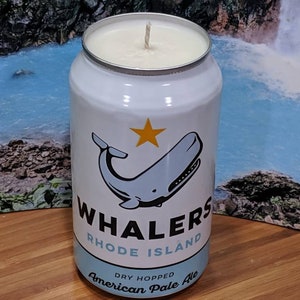 May include: A white candle in a can featuring a whale illustration and the text "Whalers Rhode Island American Pale Ale." The candle has a silver rim and a white wax top. The can design includes a blue whale and a yellow star.