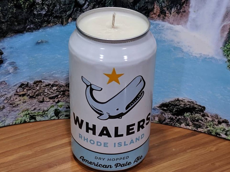 May include: A white candle in a can featuring a whale illustration and the text "Whalers Rhode Island American Pale Ale." The candle has a silver rim and a white wax top. The can design includes a blue whale and a yellow star.
