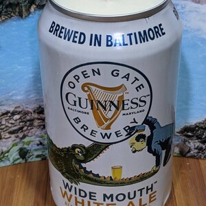 May include: A white candle in a repurposed Guinness beer can. The can features the text "BREWED IN BALTIMORE" and "OPEN GATE GUINNESS BREWER". The label also reads "WIDE MOUTH WHITE ALE" and "12 fl oz (355 ml)".