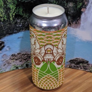 May include: A cylindrical candle in a silver can with a decorative label featuring a white and brown design on a green and orange background. The candle has a white wax and a single wick. The can is sitting on a wooden surface.