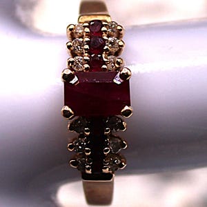 May include: A gold ring featuring a large, rectangular ruby center stone, flanked by smaller rubies and accent diamonds. The ring has a classic design with a polished gold band and is set against a white background.