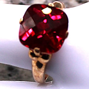 May include: A gold ring featuring a large, faceted, square-cut, deep red gemstone. The gemstone is set in a gold band with intricate detailing. The ring is displayed on a white surface, highlighting the vibrant color of the stone.