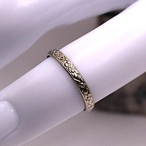 May include: A gold-colored ring with an intricate floral pattern. The ring is thin and appears to be worn on a finger. The design features detailed leaves and vines, creating a delicate and ornate appearance. The ring is likely made of metal.