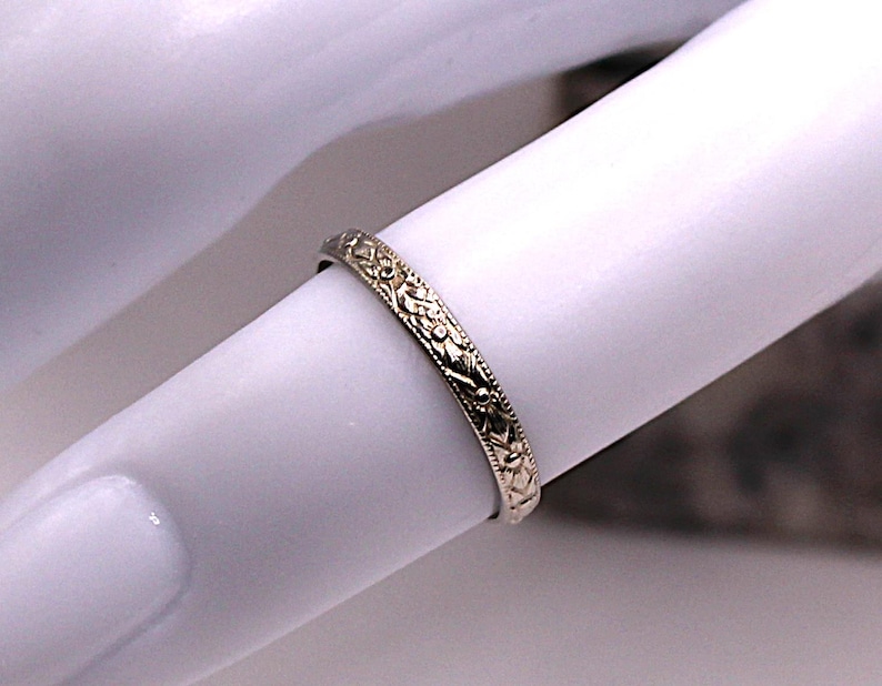 May include: A gold-colored ring with an intricate floral pattern. The ring is thin and appears to be worn on a finger. The design features detailed leaves and vines, creating a delicate and ornate appearance. The ring is likely made of metal.