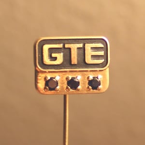 May include: A gold-toned lapel pin with the letters "GTE" in black. The pin features three small, dark gemstones below the letters. The pin is on a gold-colored stem.
