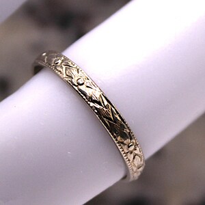 May include: A close-up of a gold-colored ring with an intricate floral and geometric pattern. The ring is displayed on a white surface, showcasing the detailed design. The ring's surface is engraved with a repeating pattern of flowers and geometric shapes.