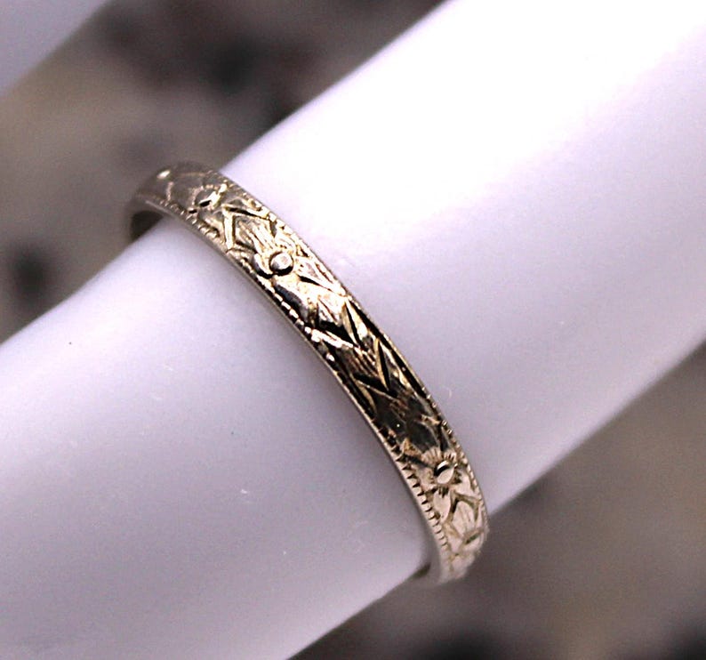 May include: A close-up of a gold-colored ring with an intricate floral and geometric pattern. The ring is displayed on a white surface, showcasing the detailed design. The ring's surface is engraved with a repeating pattern of flowers and geometric shapes.