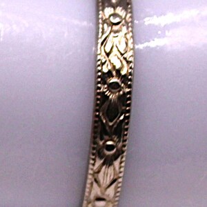 May include: A gold-colored ring with an intricate floral pattern. The ring features a repeating design of stylized flowers and leaves, with small circular accents. The band is approximately 0.2 cm wide.
