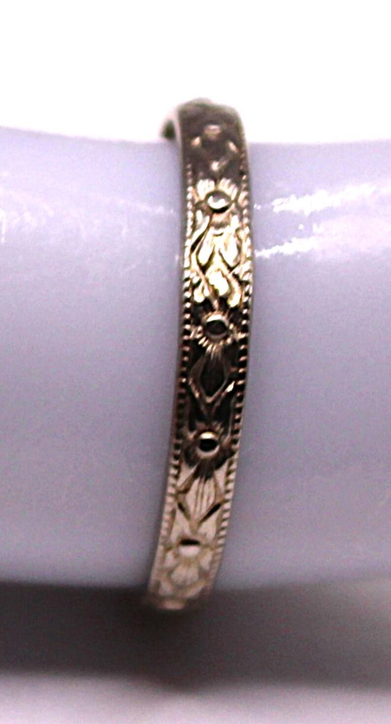 May include: A gold-colored ring with an intricate floral pattern. The ring features a repeating design of stylized flowers and leaves, with small circular accents. The band is approximately 0.2 cm wide.