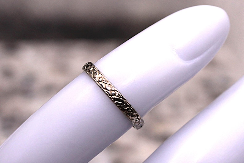 May include: A gold-colored ring with an intricate floral pattern. The ring is thin and sits on a white surface. The ring's design is detailed and appears to be vintage or antique.