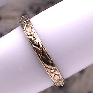 May include: Close-up of a gold-colored ring with an intricate floral and geometric pattern. The ring is set against a white background, showcasing the detailed design. The ring's band is approximately 2 mm wide.