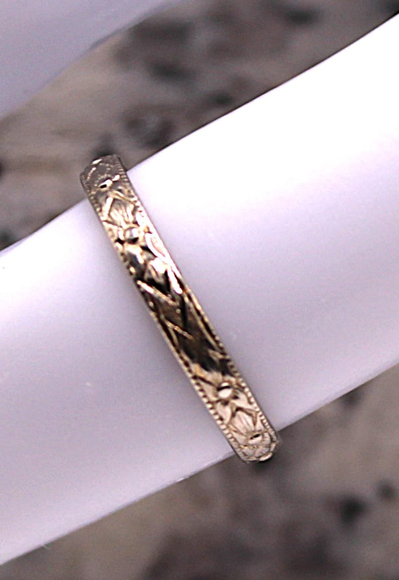 May include: Close-up of a gold-colored ring with an intricate floral and geometric pattern. The ring is set against a white background, showcasing the detailed design. The ring's band is approximately 2 mm wide.