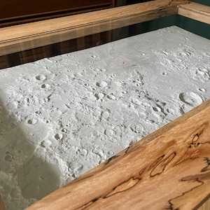May include: A detailed model of the moon's surface, with craters and other features, displayed in a wooden frame with a glass top.