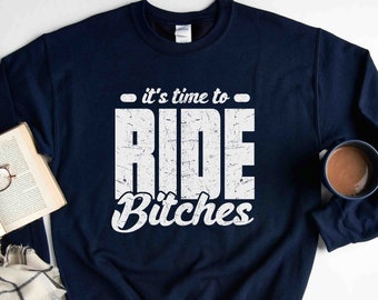 Op de afbeelding: Marineblauwe sweatshirt met de tekst "it's time to RIDE Bitches" in verweerde witte letters. De afbeelding bevat een boek, een bril, een geruite sjaal en een mok koffie.