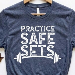 Könnte beinhalten: Ein marineblaues T-Shirt mit dem weißen Text "PRACTICE SAFE SETS" und einer Hantel-Grafik. Das T-Shirt wird auf einem Holzbügel präsentiert, daneben liegt eine braune Sonnenbrille. Das T-Shirt ist unten geknotet.