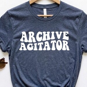 May include: A heather navy blue short-sleeved t-shirt featuring the white text ARCHIVE AGITATOR in a wavy, retro-inspired font. The shirt is displayed on a wooden hanger and is accompanied by a pair of brown tortoiseshell sunglasses.