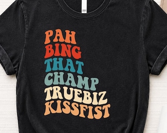 May include: Black t-shirt featuring the text "PAH BING THAT CHAMP TRUEBIZ KISSFIST" in a retro font. The text is in orange, red, blue, teal, and cream. The shirt has a crew neck and short sleeves.