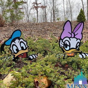 May include: A pair of cartoon character yard decorations, Donald Duck and Daisy Duck, peeking out from behind a bush. Donald Duck is wearing a blue sailor hat and Daisy Duck is wearing a pink bow.