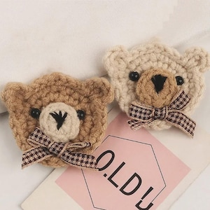 May include: Two crocheted bear appliques, one in medium brown and one in cream, each featuring black bead eyes, a stitched black nose, and a brown and white checkered ribbon bow tied at the neck, positioned above a pink card with the text OLD.