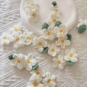 Könnte beinhalten: Eine Sammlung handgefertigter Häkelblumen mit weißen Blütenblättern und gelben Mitten, akzentuiert durch kleine grüne Blätter. Diese einzelnen Blumenapplikationen sind aus Garn gefertigt und in einem Streumuster auf einer weißen Fläche angeordnet.