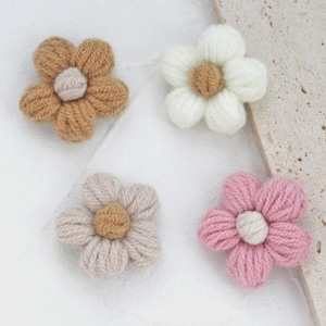 May include: Four handmade flower appliques in shades of brown, white, pink, and beige. Each flower has a central, textured detail. The petals are made of yarn, giving them a soft, tactile appearance. These decorative elements are ideal for craft projects.