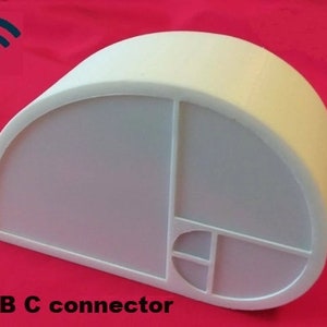 Puede incluir: Altavoz inalámbrico blanco, semicircular, con un conector USB-C. El altavoz tiene un diseño geométrico en la parte delantera.