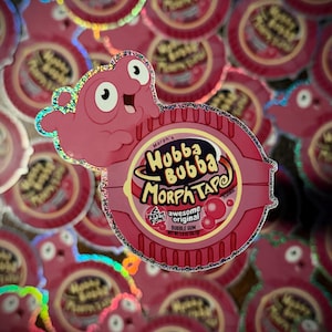 May include: A holographic sticker featuring a pink cartoon character leaning over a round Hubba Bubba Morph Tape bubble gum container. The label text reads Morph's Hubba Bubba Morph Tape, 6 feet of fun, awesome original, and bubble gum.