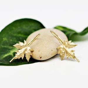 May include: A pair of gold leaf dangle earrings. The earrings are shaped like maple leaves and are hanging from a small hook.