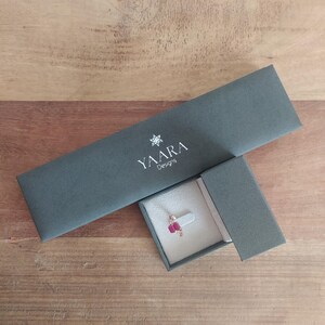 Puede incluir: Una caja de joyer&iacute;a gris oscuro con el texto "YAARA Designs" en la tapa. La caja est&aacute; abierta y un peque&ntilde;o anillo con una piedra preciosa roja est&aacute; dentro.