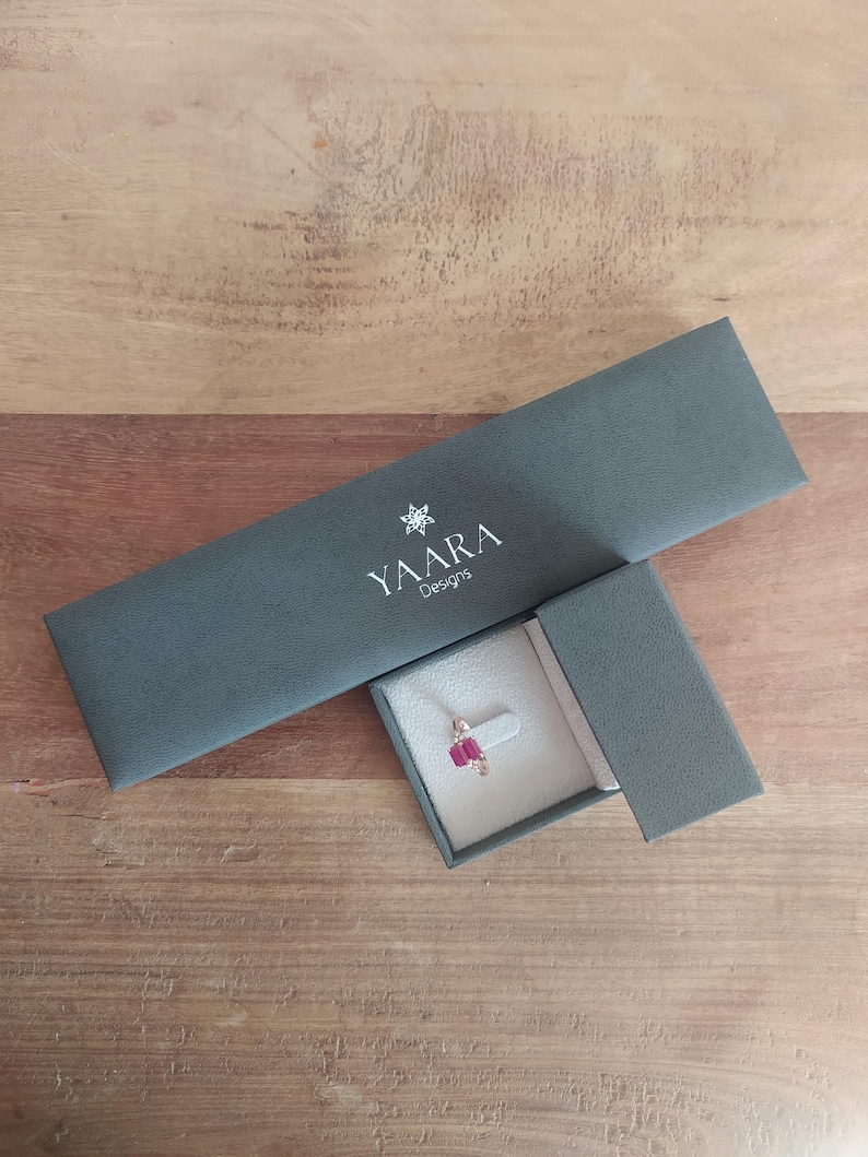 Puede incluir: Una caja de joyer&iacute;a gris oscuro con el texto "YAARA Designs" en la tapa. La caja est&aacute; abierta y un peque&ntilde;o anillo con una piedra preciosa roja est&aacute; dentro.