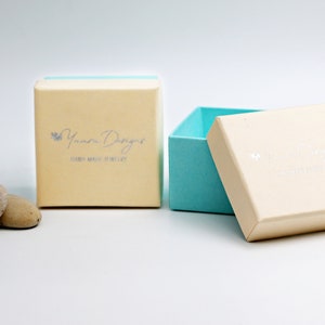 May include: A light blue and cream colored jewelry box with the text "Yumna Designs" and "Hand Made Jewelry" on the lid. The box is open and the inside is light blue.