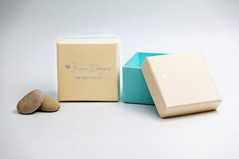 May include: A light blue and cream colored jewelry box with the text "Yumna Designs" and "Hand Made Jewelry" on the lid. The box is open and the inside is light blue.