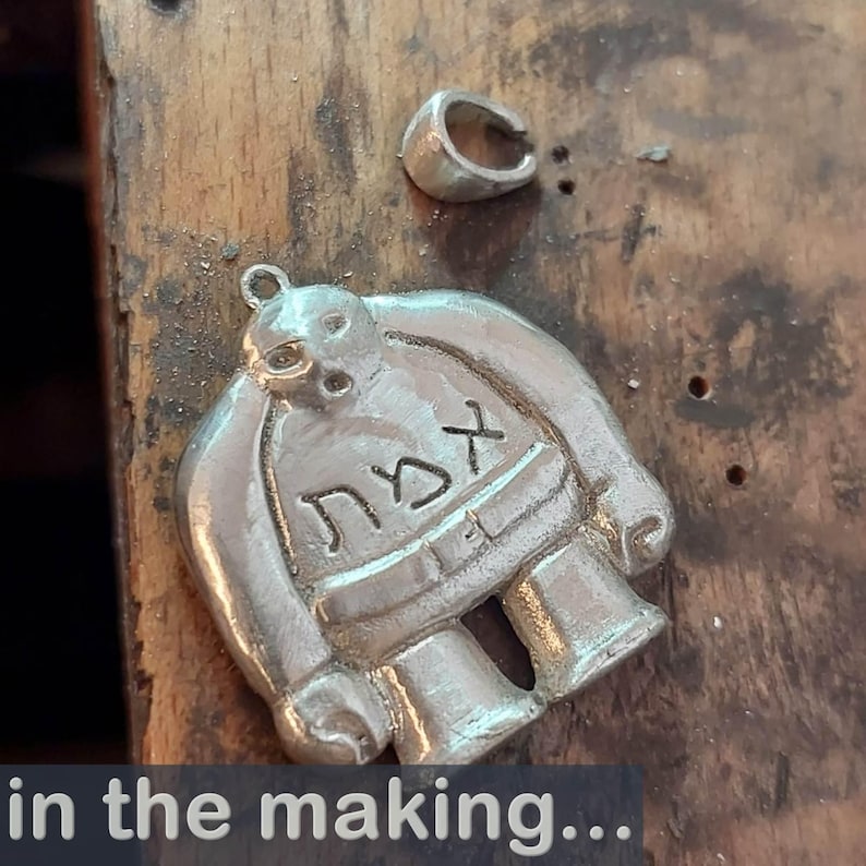 Puede incluir: Un colgante de plata con forma de personaje de dibujos animados con la palabra hebrea "אמת" (verdad) inscrita en su pecho. El colgante se est&aacute; haciendo y est&aacute; sobre una superficie de madera.