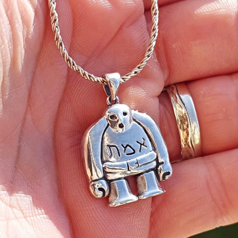 Puede incluir: Un collar de plata con un colgante en forma de una peque&ntilde;a figura de dibujos animados. La figura tiene una cabeza redonda con una cara, un cuerpo con brazos y piernas, y la palabra "אצח" escrita en su pecho.