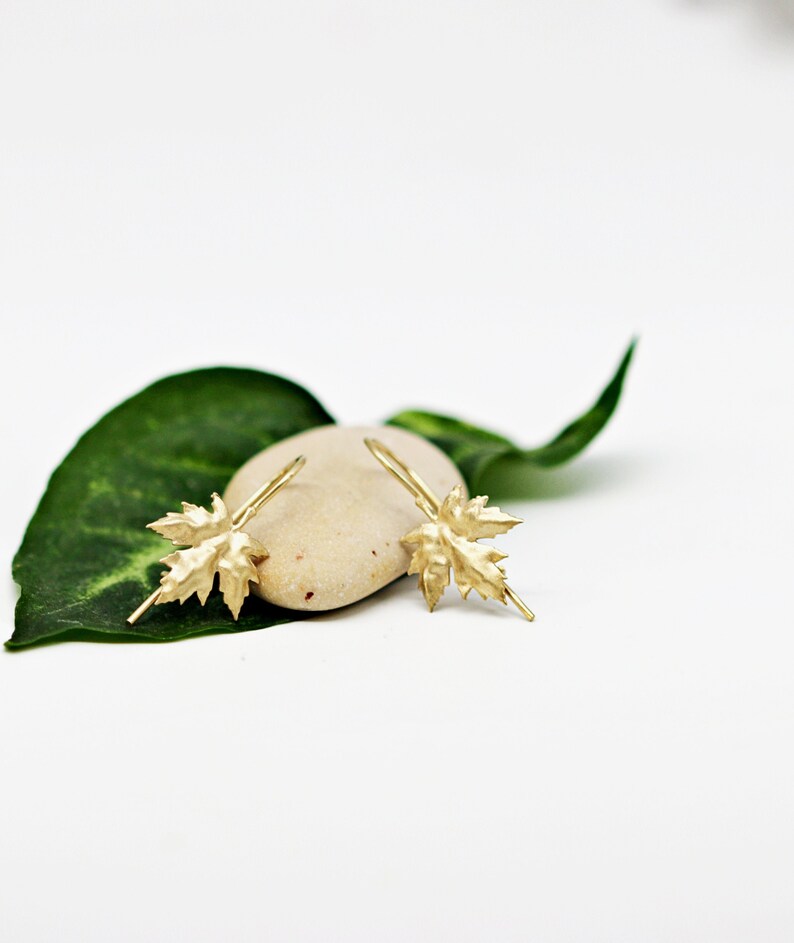 May include: A pair of gold leaf dangle earrings. The earrings are shaped like maple leaves and are hanging from a small hook.