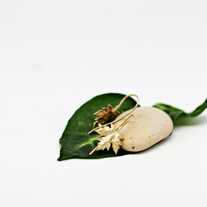 May include: Gold leaf earrings with a delicate design, resting on a small, light brown stone and a green leaf.