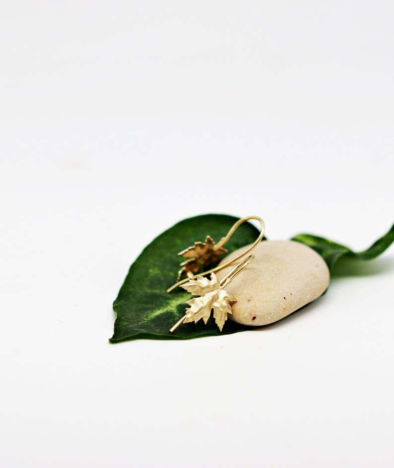May include: Gold leaf earrings with a delicate design, resting on a small, light brown stone and a green leaf.