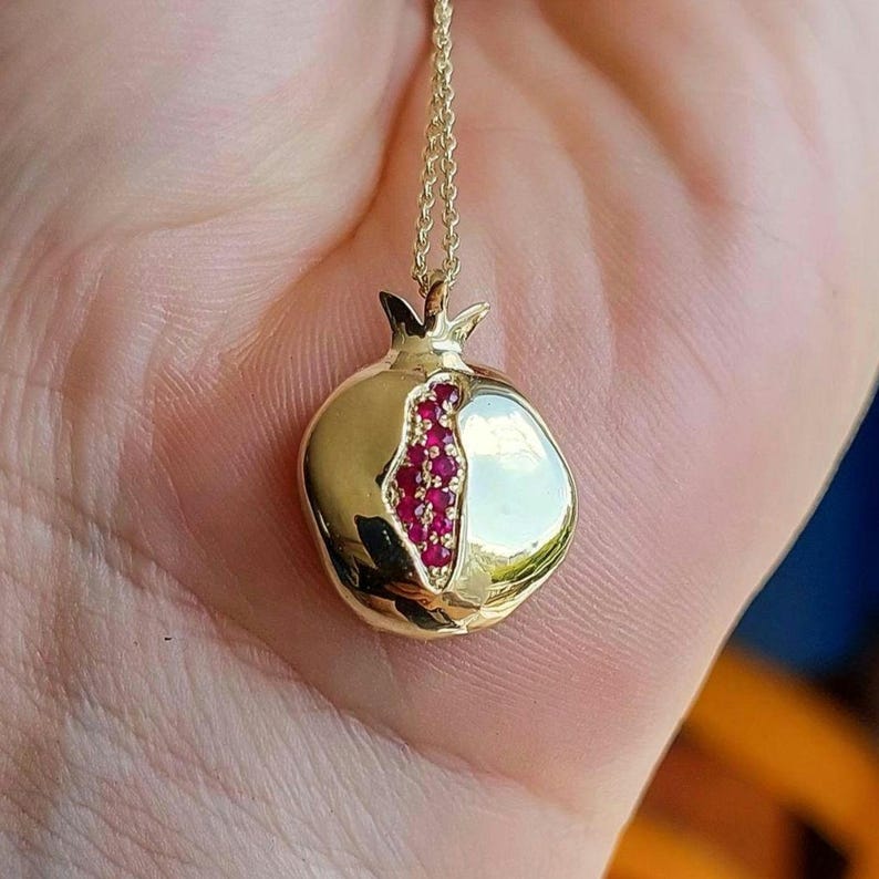 Puede incluir: Un collar de oro con un colgante de granada. El colgante est&aacute; hecho de oro y tiene piedras de rub&iacute; rojo engastadas en forma de semilla de granada.
