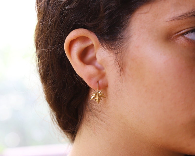 May include: Gold leaf earrings with a delicate design. The earrings are small and dangle from the earlobe.