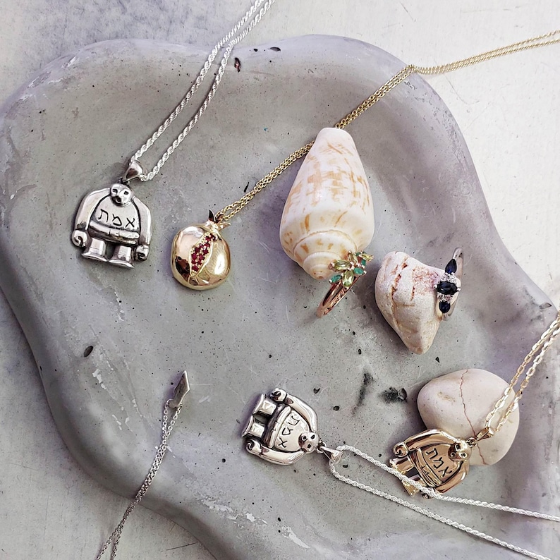 Puede incluir: Una colecci&oacute;n de collares de plata y oro con colgantes que presentan texto hebreo, una granada y una concha. Los collares se muestran sobre una superficie gris texturizada.