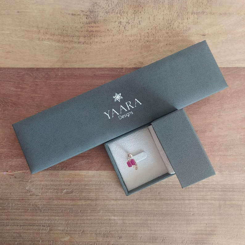 Puede incluir: Una caja de joyer&iacute;a gris con el texto "YAARA Designs" en la tapa. La caja est&aacute; abierta y un anillo con una piedra preciosa roja est&aacute; dentro.