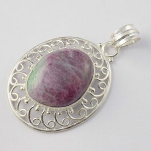 May include: Oval pendant with a polished, multi-colored gemstone, primarily ruby red with green and white hues. The stone is set in a decorative, openwork silver-tone metal frame with a bail for a chain.
