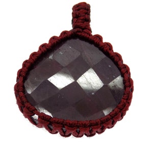 May include: A faceted, dark burgundy gemstone pendant with a teardrop shape. The stone is encased in a macrame-style border and a loop for hanging, all in a matching burgundy color. The pendant is set against a white background.