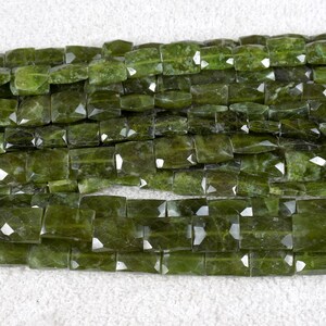 May include: Close-up of a strand of faceted, rectangular green gemstone beads. The beads have a polished, reflective surface and are arranged in neat rows. The color is a deep, olive green.