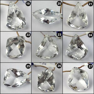 May include: A collection of clear, faceted gemstone pendants in various shapes, including teardrop, marquise, and square. Each pendant is strung on a gold-colored wire, showcasing the internal facets and clarity of the stones. The pendants are displayed against a white background.