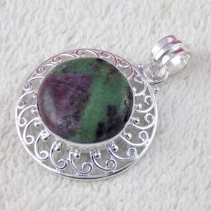 May include: A silver pendant with a round, polished stone featuring a mix of deep red and green hues. The stone is set within a decorative, openwork silver frame. The pendant has a loop for a chain.