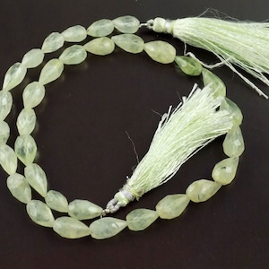 May include: A necklace featuring faceted, pear-shaped, pale green prehnite beads. The necklace is accented with two light green tassels and silver-colored metal findings. The beads are strung together, creating a delicate and elegant design.