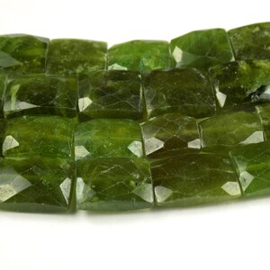 May include: Close-up of a strand of faceted, rectangular green gemstones. The stones have a deep, olive-green color with multiple facets that catch the light. The gemstones are arranged closely together, suggesting they are strung for jewelry making.